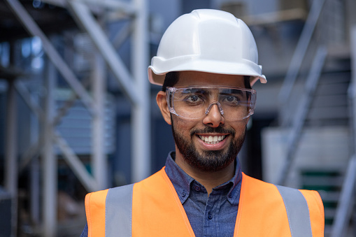 Close-up portrait of young engineer in hard hat and vest, man smiling and looking at camera, worker in factory plant, and logistics warehouse.