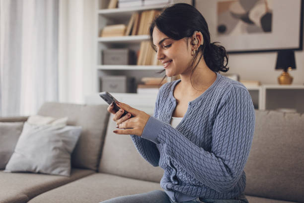 istock Young beautiful woman using smart phone while wearing her hearing aid at home 1805613141