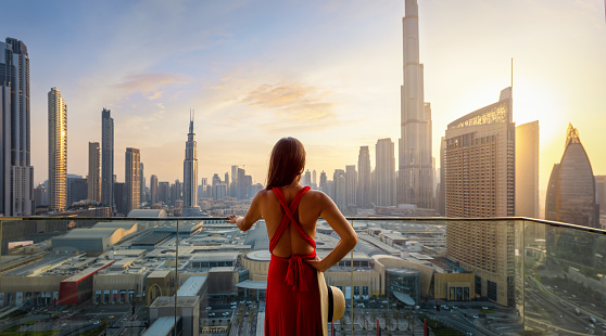 A beautiful luxury woman in a red dress enjoys the sunset view over the modern skyline of Dubai city, UAE