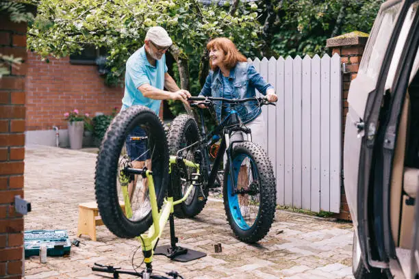 Active senior couple, checks the tires and gears of their fatbikes, preparing for an adventurous vacation with their trusty campervan parked near them.