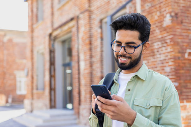 istock Portrait of young successful student outside university campus, man smiling and using app on phone, typing message and browsing social media, indian man holding phone with backpack 1805644714