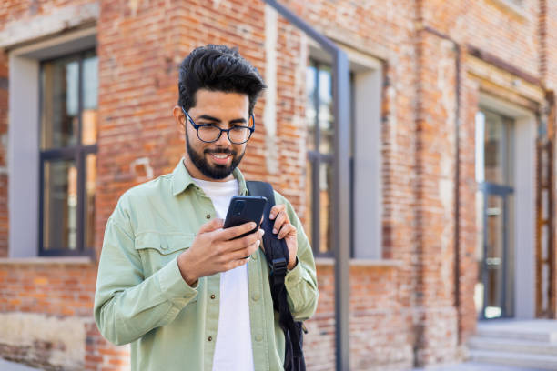 istock Portrait of young successful student outside university campus, man smiling and using app on phone, typing message and browsing social media, indian man holding phone with backpack 1805644718