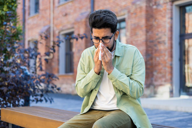 istock A man sneezes while sitting on a bench outside an office building, a young businessman has a runny nose and allergies, sick 1805649864