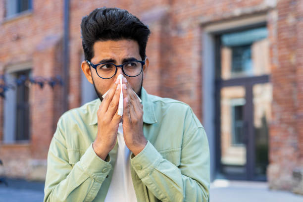 istock A man sneezes while sitting on a bench outside an office building, a young businessman has a runny nose and allergies, sick 1805649865