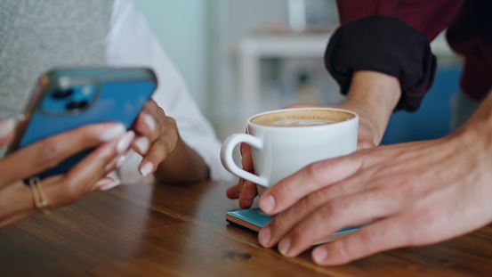 Barista putting cup coffee on table to woman sitting with smartphone closeup.