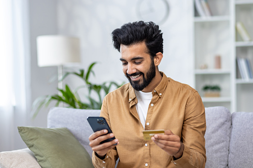 Young joyful man sitting on sofa at home, using application on phone for online shopping in online store, Indian man smiling contentedly holding bank credit card.