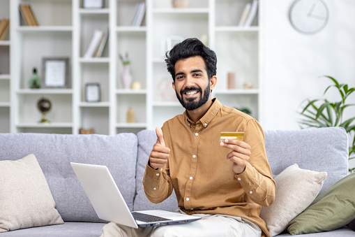 Happy Indian man sitting at home on sofa with laptop on lap, holding card in hands, very happy about online shopping, online payment, showing thumb up, smiling, looking at camera.