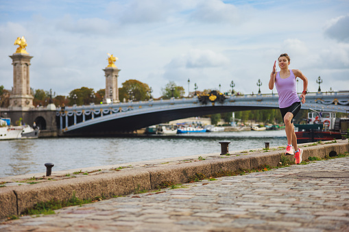 Active young woman in sports clothing jogging on stone paved pier on Paris waterfront, cloudy summer day by Seine river