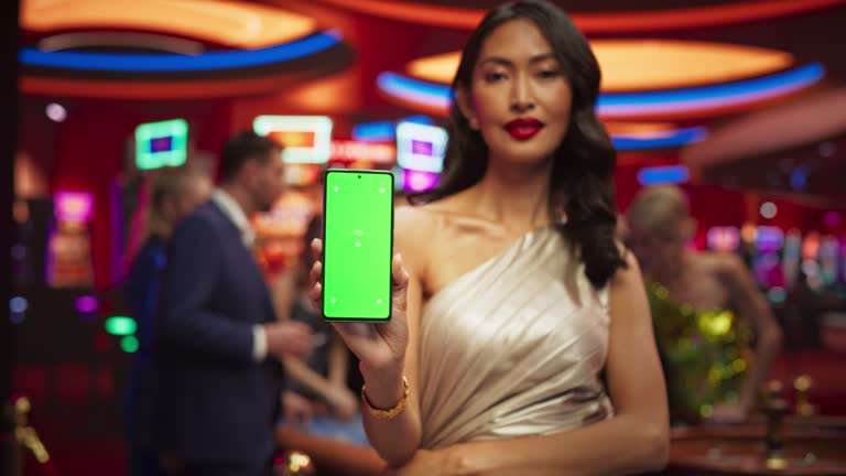 Advertising Template with an Asian Female Showing a Smartphone Device with a Green Screen Mock Up Display with Trackers. Professional Beautiful Model in a Casino with People Gambling in the Background