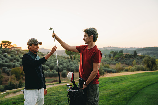 Caddy is giving a club to the golfer on green. Sunset time on the golf course.