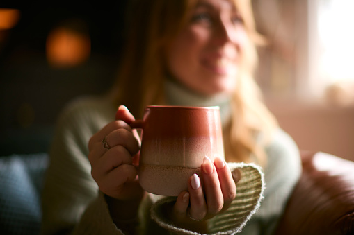 Smiling Woman At Home Wearing Winter Jumper With Warming Hot Drink Of Tea Or Coffee In Cup Or Mug