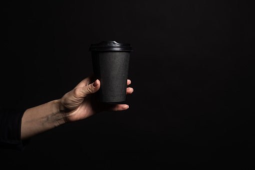 Black paper cup of coffee in hand isolated on a black background. Mock-up