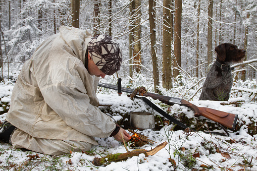 Winter forest. A hunter lights a fire under a hanging pot. He is wearing white camouflage. A hunting dog is sitting nearby.