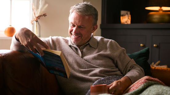 Relaxed elderly man sitting on a chair reading a book and drinking coffee at home