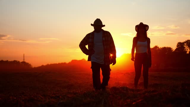 Silhouette of two farmers in straw hat, walking across plowed land field in red sun rays sunset. Couple do family agribusiness. Partners walk and inspect their rural territories lands. Countryside.
