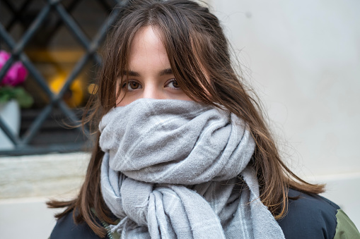 Portrait of beautiful teenage girl with brown hair, wearing winter jacket and scarf over her mouth and nose, showing only her eyes, looking at camera