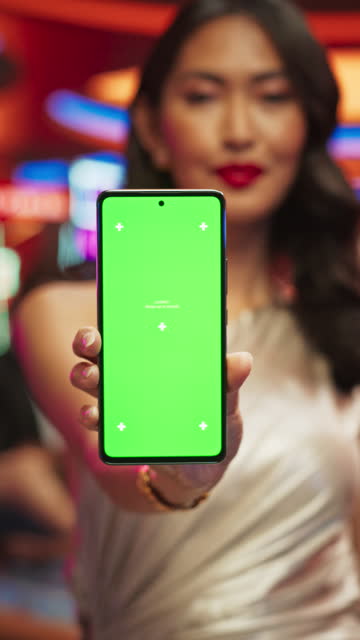 Vertical Screen: Advertising Template with an Asian Female Showing a Smartphone with a Green Screen Mock Up Display. Professional Beautiful Model in a Casino with People Gambling in the Background