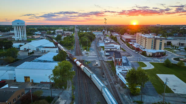istock Golden sun setting on horizon with hotel and water tower split by train on railroad tracks aerial 1805802217