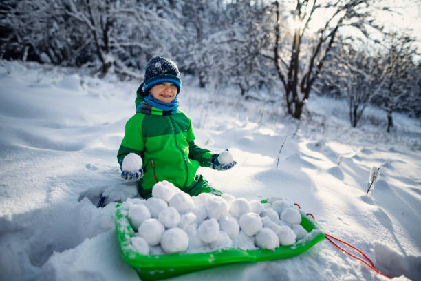 Little boy preparing snowballs for a snowball fight in the forest. The boy has prepared a sled full of snowballs in front of him.\nShot with Nikon D850