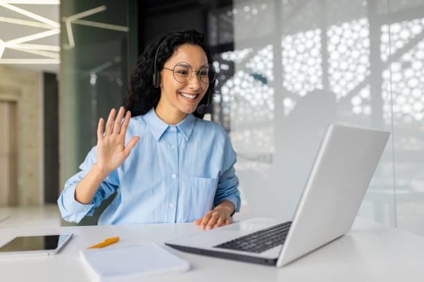 istock Young female online support worker using headset phone for video call, consulting customer consumers remotely, businesswoman working inside office with laptop, waving at camera saying hello 1805848671