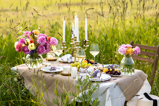 Beautiful romantic outdoor wedding decor in a field. Table decorated with ranunculi flowers. Wineglasses with white wine. Sunset, summer, golden hour. Perfect surprise date for loving couple