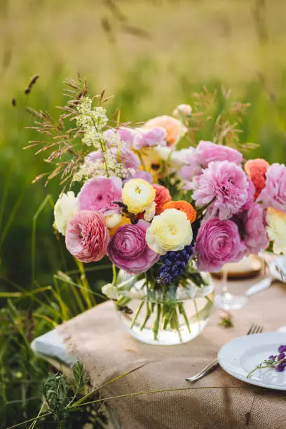 Beautiful romantic outdoor wedding decor in a field. Table decorated with ranunculi flowers. Wineglasses with white wine. Sunset, summer, golden hour. Perfect surprise date for loving couple