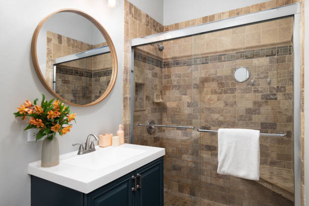 A bathroom with a blue cabinet and a stone tiled shower. A bathroom with a blue cabinet wooden circular mirror above a white sink, and a shower with brown stone tiles. renovation stock pictures, royalty-free photos & images