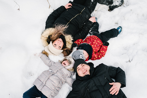 Happy family lying down in the snow in park. Top view. Father, mother, children walking snowy mountains, time together. Winter holidays. Mom, dad hugging son, daughter lying in snowy winter forest.