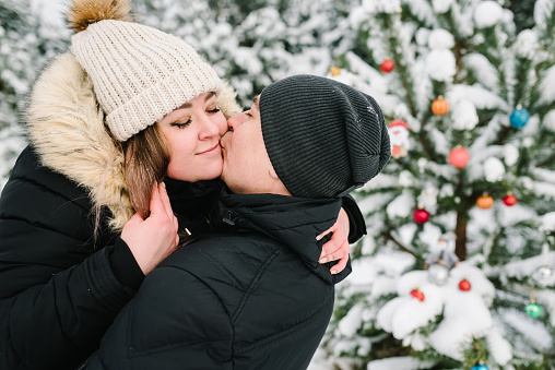 Man kisses woman standing near decorated Christmas tree in park. Couple smiling having fun, hugging in snowy winter forest. Young guy and girl in winter wear enjoying snowfall. Happy winter holidays.