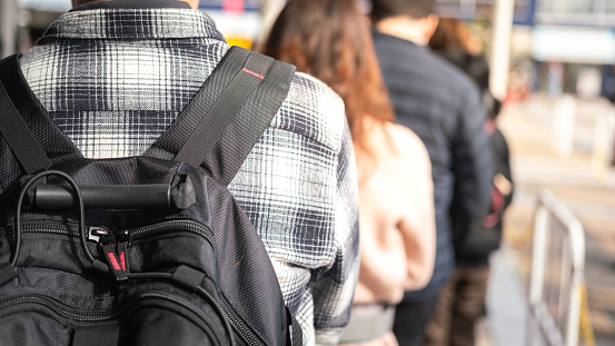 Close-up at back of a man with backpack bag during queue up and waiting to buy a attractive event ticket, with blurred background of many people in queue.