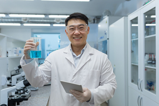 Portrait of a young Asian male student studying in the laboratory. Standing holding a flask of liquid and a tablet, pointing to the camera and smiling.