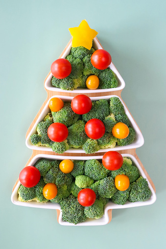 Stock photo showing close-up view of Christmas party food buffet platter. Christmas tree shaped wood and china tray filled with broccoli florets decorated with baubles made from red cherry tomatoes and pieces of yellow bell pepper displayed on a light green background.