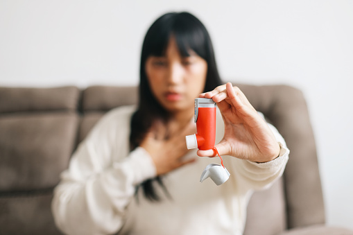 Woman showing asthma inhaler while holding chest in house