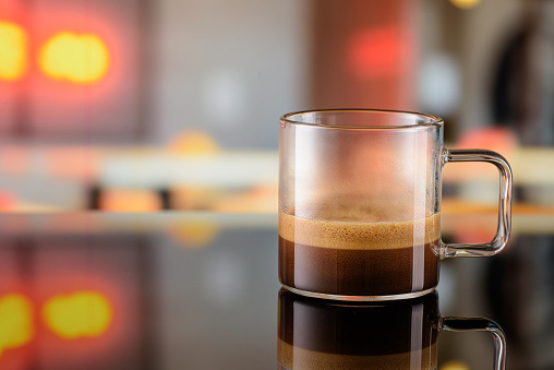 Glass transparent cup of ristretto coffee on the bar counter with blurred lights background. Copy space for your text.
