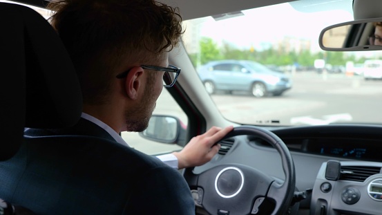 Young businessman wearing glasses and driving around the city at his car. Back view. Transport, business and people concept. Slow motion