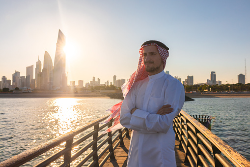 Sheik wearing a robe walking on a bridge with the cityscape in the background.