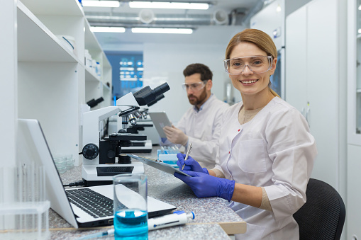 Portrait of female scientist laboratory assistant, researcher working inside medical laboratory with microscope, looking at camera and smiling while sitting, female worker in white medical coat.