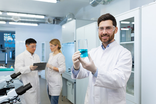 Retrato de un joven químico de pie en un laboratorio y sosteniendo un matraz con una sustancia azul desconocida en sus manos y sonriendo mirando a la cámara photo