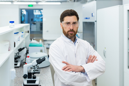 Portrait of a serious young male scientist, medical worker, chemist standing in a laboratory in a white coat and protective glasses, looking at the camera with his arms crossed.