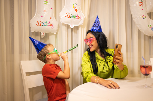 Excited Caucasian boy, celebrating his birthday with his mother/aunt/nanny or grandmother, while she taking a selfie while he blowing a party horn blower