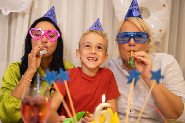 niño caucásico, celebrando su 6º cumpleaños con sus madres, en su casa mientras sopla una bocina de fiesta - ahijado fotografías e imágenes de stock