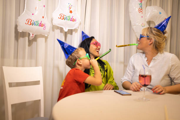 niño caucásico celebrando su cumpleaños mientras sopla una bocina de fiesta con su madre y su tía - ahijado fotografías e imágenes de stock
