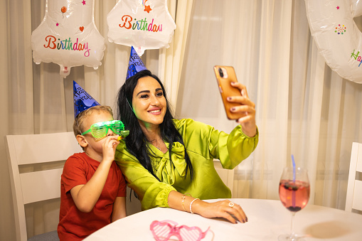 Excited Caucasian boy, celebrating his birthday with his mother/aunt/nanny or grandmother, while she taking a selfie while he blowing a party horn blower