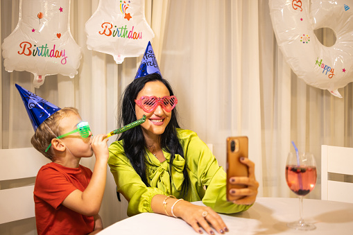 Excited Caucasian boy, celebrating his birthday with his mother/aunt/nanny or grandmother, while she taking a selfie while he blowing a party horn blower
