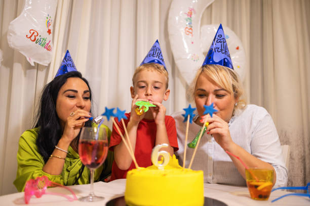 niño caucásico feliz celebrando su 6º cumpleaños con sus madres, mientras sopla un soplador de bocina de fiesta - ahijado fotografías e imágenes de stock
