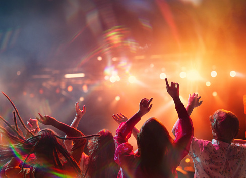large group of people having fun, singing and dancing raising hands on concert of favorite artists in neon light, spotlights near stage. Concept of party, festivals, concerts, music and dance.