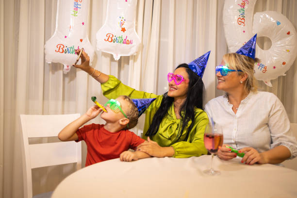 niño caucásico celebrando su cumpleaños mientras sopla un soplador de bocina de fiesta con su madre y su niñera - ahijado fotografías e imágenes de stock
