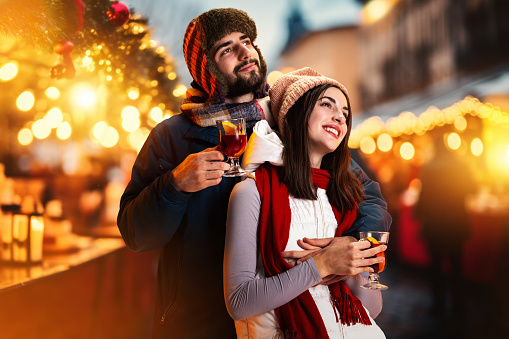 Young romantic couple having fun outdoors in winter fair and hugs looking to sky on festive salute, fireworks in New Year night. Concept of national traditions, winter holidays, fashion, festivities