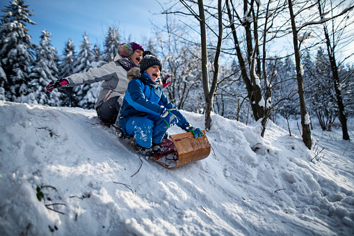 Little boy and his sister are sledding together on retro style toboggan in the beautiful winter forest. Kids are 12 and 8 years old. They are shouting while riding down the small hill.\nShot with Nikon D850