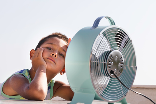 Overheated little boy is sitting in front of electric fan trying to cool down. Climate change concept.